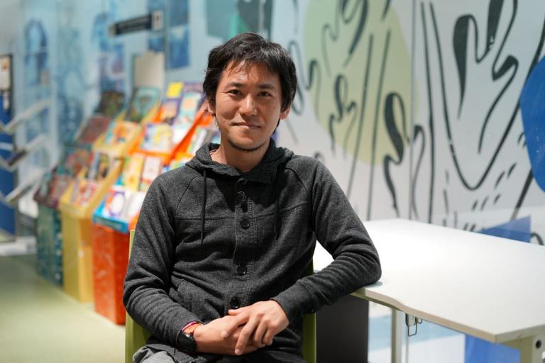 Ritsu is seated and looking at the camera and smiling. The background is some colourful children's books and a mural painted on the wall
