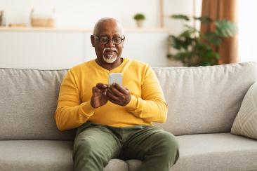 An elderly black male in a yellow sweater using a smartphone while sitting on a sofa