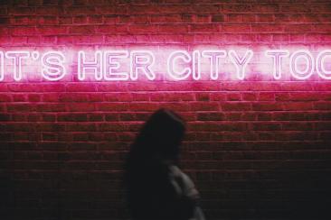 'It's her City Too' illuminated in LED lights on a brick wall at nighttime with a woman walking past. 