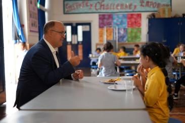 Cllr Adam Hug is facing a school child who is wearing a yellow t-shirt and eating a school meal