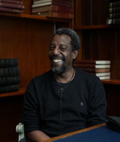 A member of staff sat in Mayfair library in front of some book shelves. He is smiling at the camera.