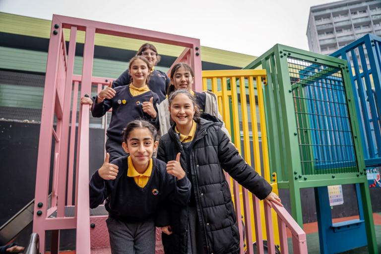 A group of children posing with thumbs up on the climbing frame's steps. The climbing frame and slide structure is multicoloured split into pink, yellow, green and blue segments.  