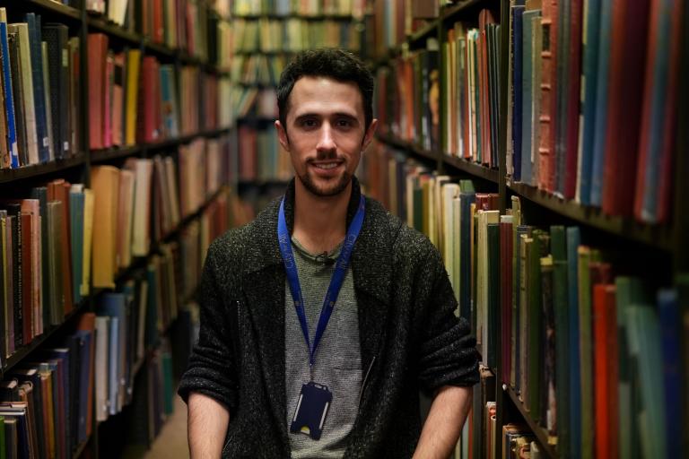 Reference Library staff member Nick standing in between a row of books, the books are all different shapes, colours and sizes.