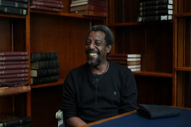 Andrew, sitting in a chair, facing the camera and smiling. Behind him are some old bound books on wooden shelves.