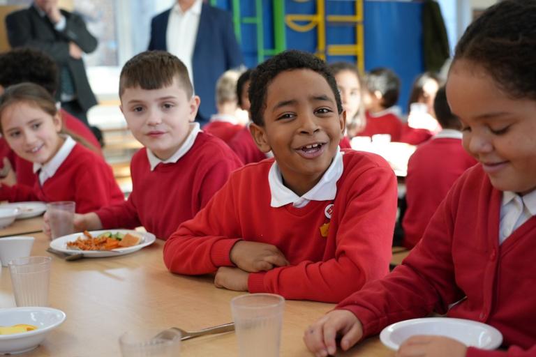 Children sat at a long wooden table having school lunch. 