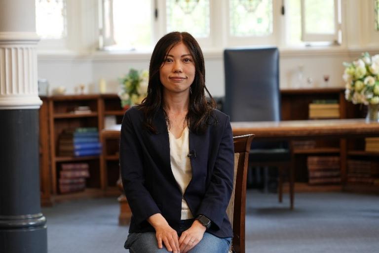 Hitomi is facing the camera and is sat on a chair in a library