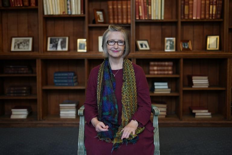 Kate is facing the camera, sitting on a chair and behind her is a wall of books in a library