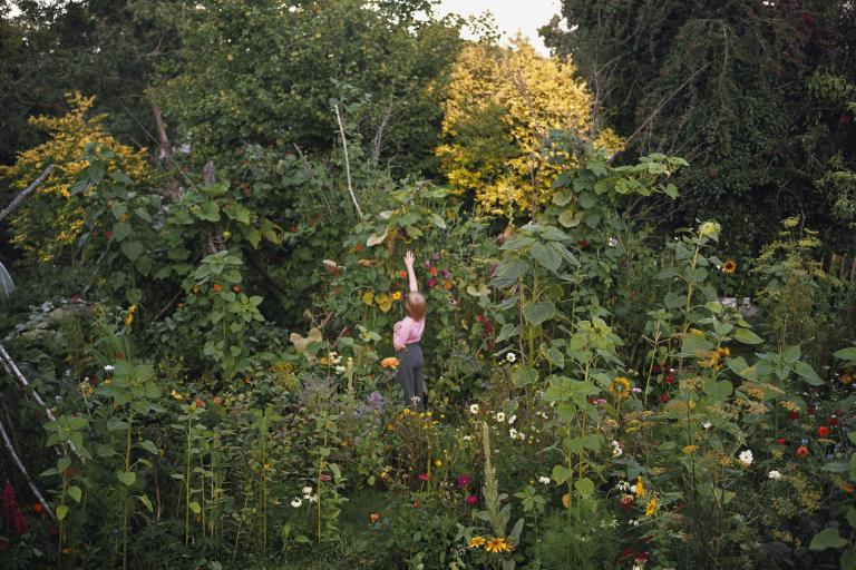 A photo of a woman in a garden reaching up to grab a leaf.