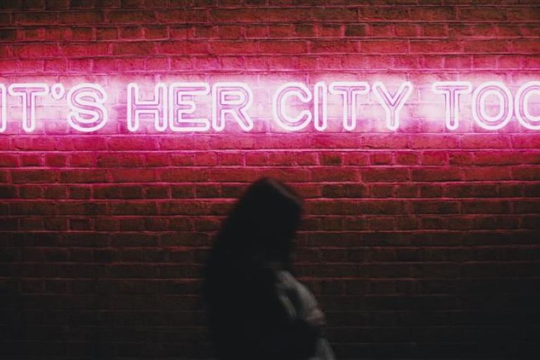 'It's her City Too' illuminated in LED lights on a brick wall at nighttime with a woman walking past. 