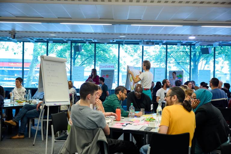 People sat around tables and one person in the middle putting a piece of paper on a board