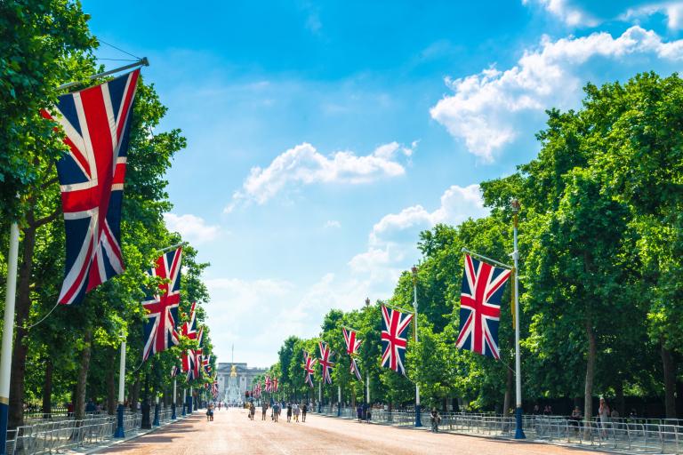 The Mall on a sunny day with some tourists walking down the road, with Union Jacks and green trees flanking either side of the road and Buckingham Palace can be seen in the distance